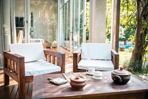 Modern patio setup with wooden armchairs, white cushions, and coffee table outside glass-walled home.