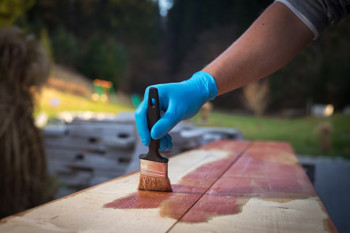 A person with blue gloves uses a brush to apply wood stain on wooden planks outdoors.