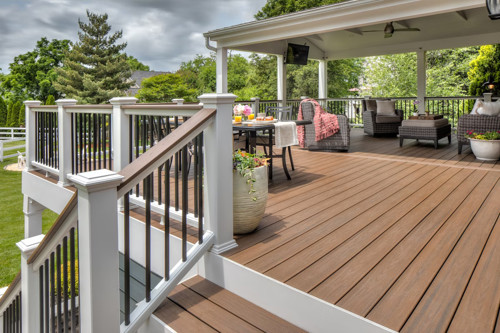 Covered brown composite deck with patio furniture, dining table, and white railing overlooking a backyard.