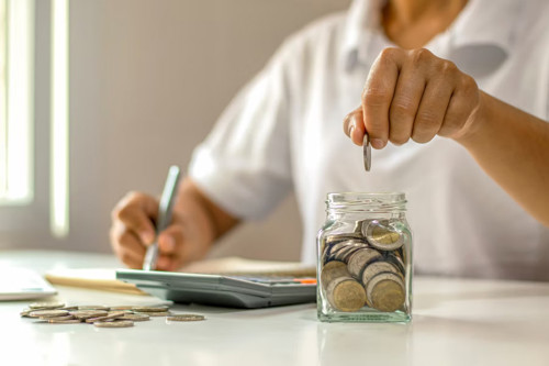 Close-up of person budgeting by placing coins in a jar with calculator and notepad on desk.